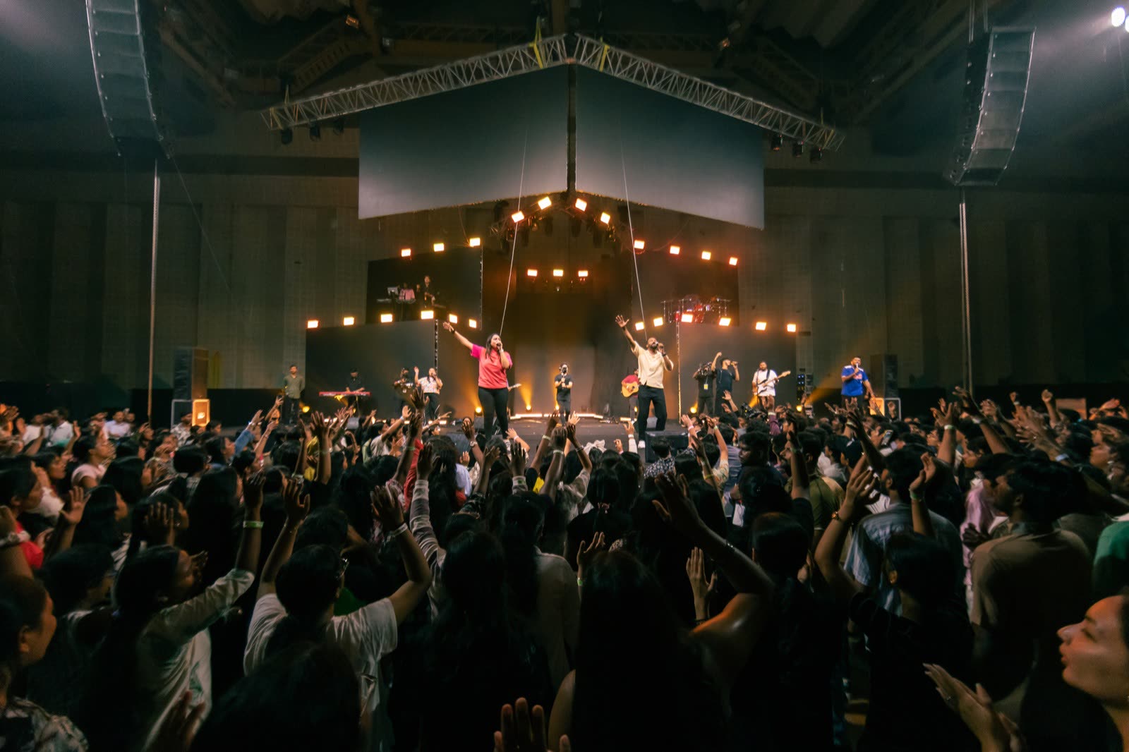Crowd with hands raised facing a worship band on stage under warm lights