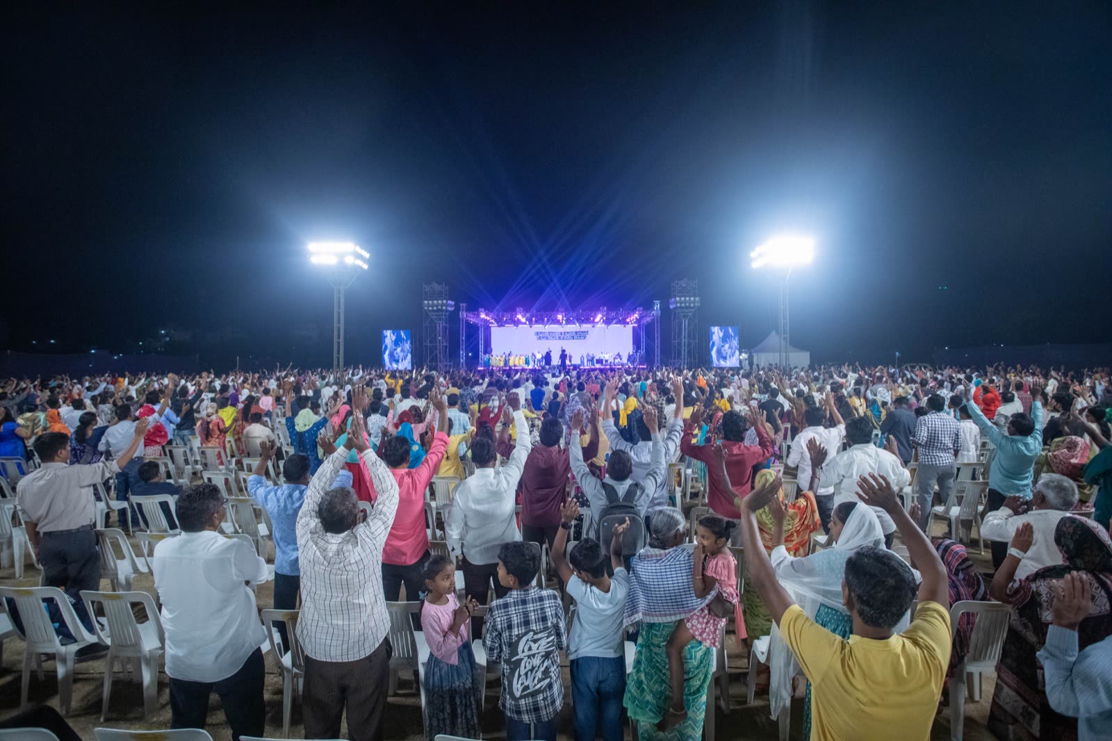 Outdoor night gathering with floodlights and a crowd facing a distant stage