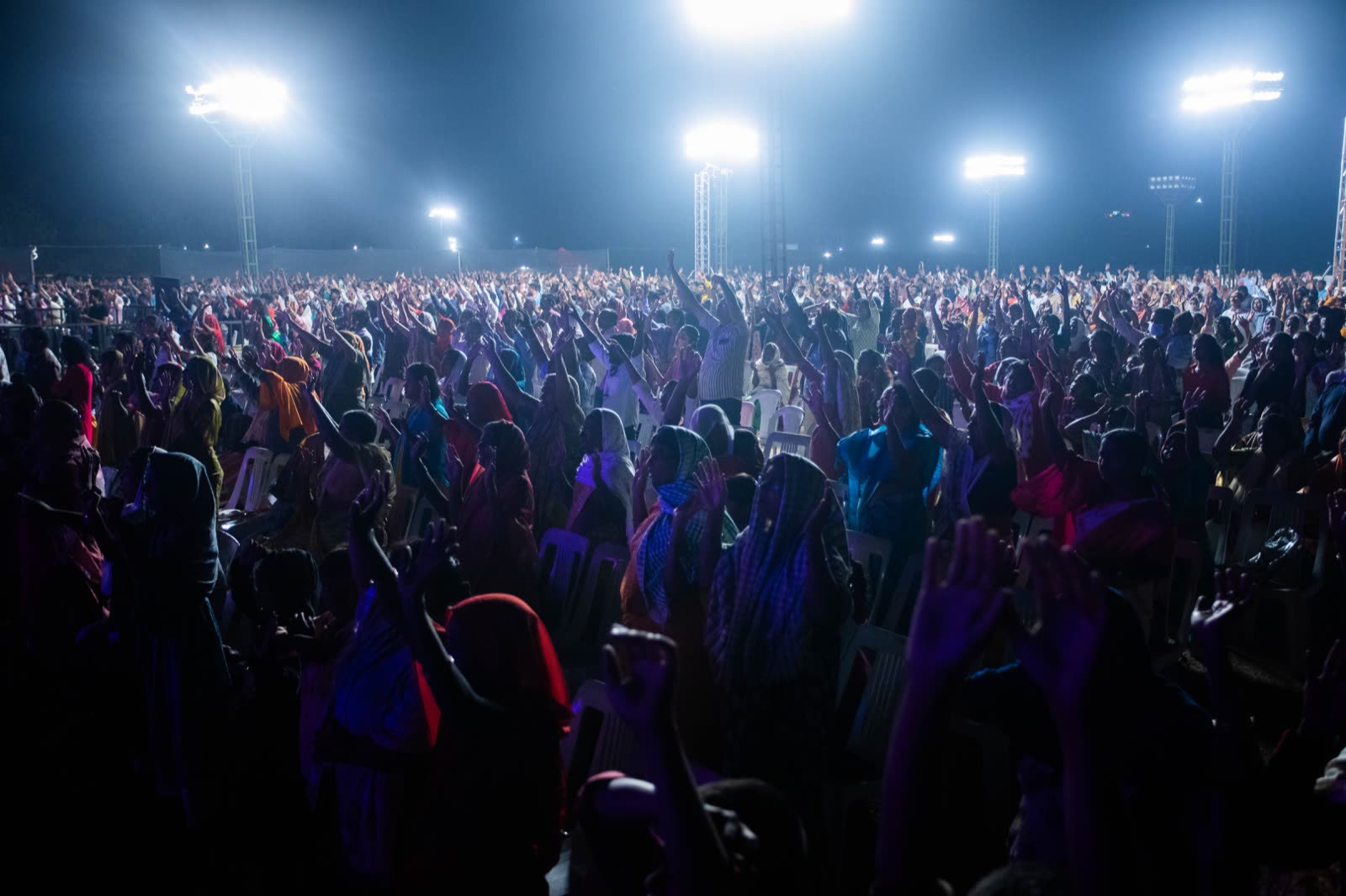 Wide night shot of a large outdoor crowd with arms raised under stadium lights