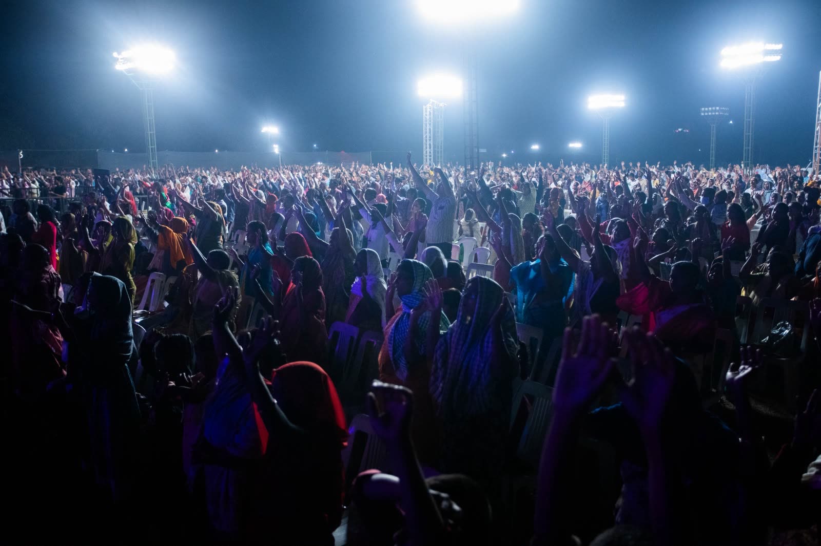 Nighttime outdoor congregation silhouetted under bright floodlight towers
